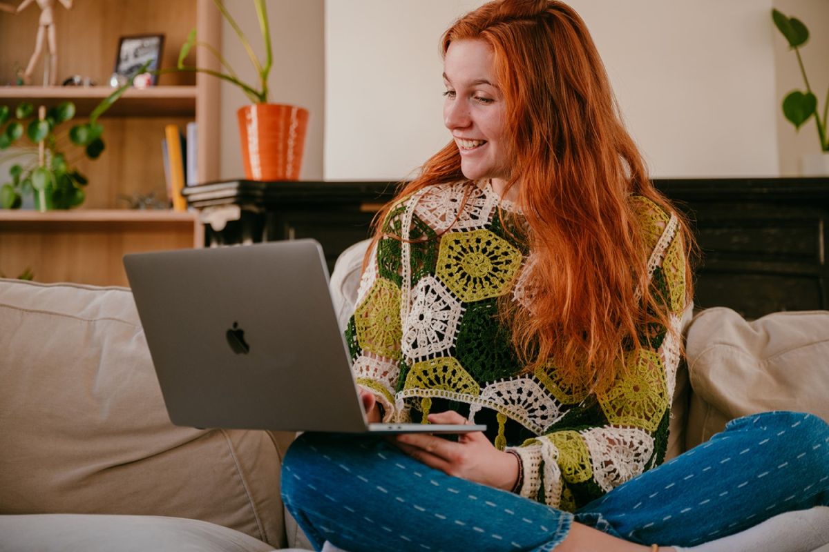 Yasmine from Learn Dutch with Yas sitting in a sofa with a laptop on her knee and smiling at the screen